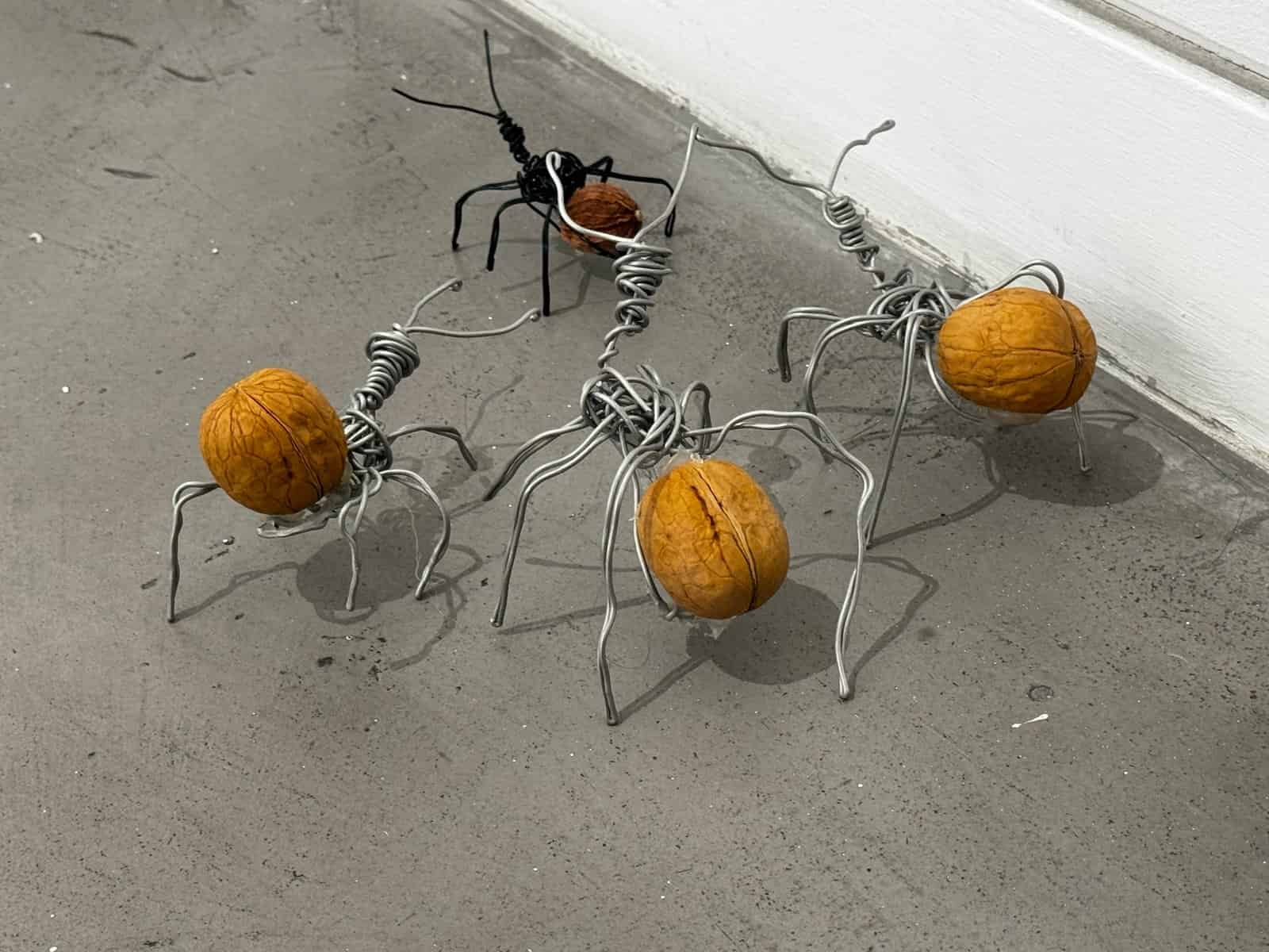 A group of hand-formed black and silver wire ants carry walnuts across a concrete gallery floor beside a white wall.