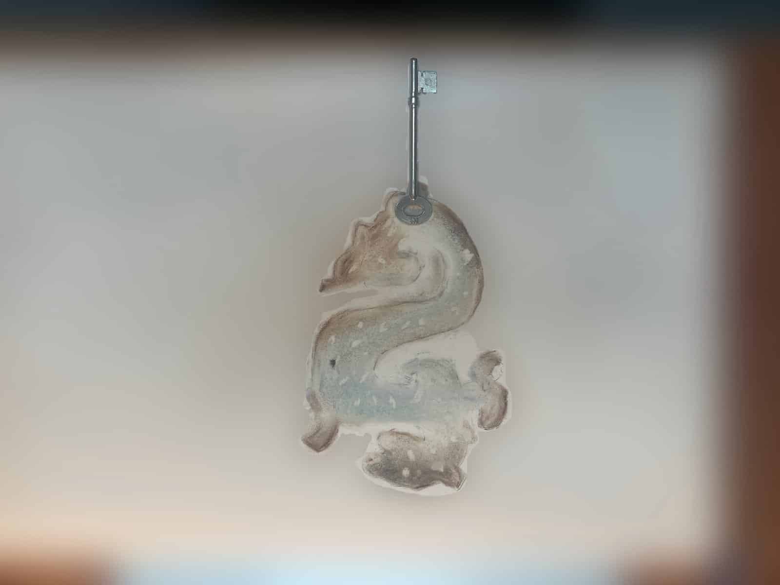 A pale translucent, heraldic-looking sculptural form hangs from an old metal key on a slender stand against a softly lit background.