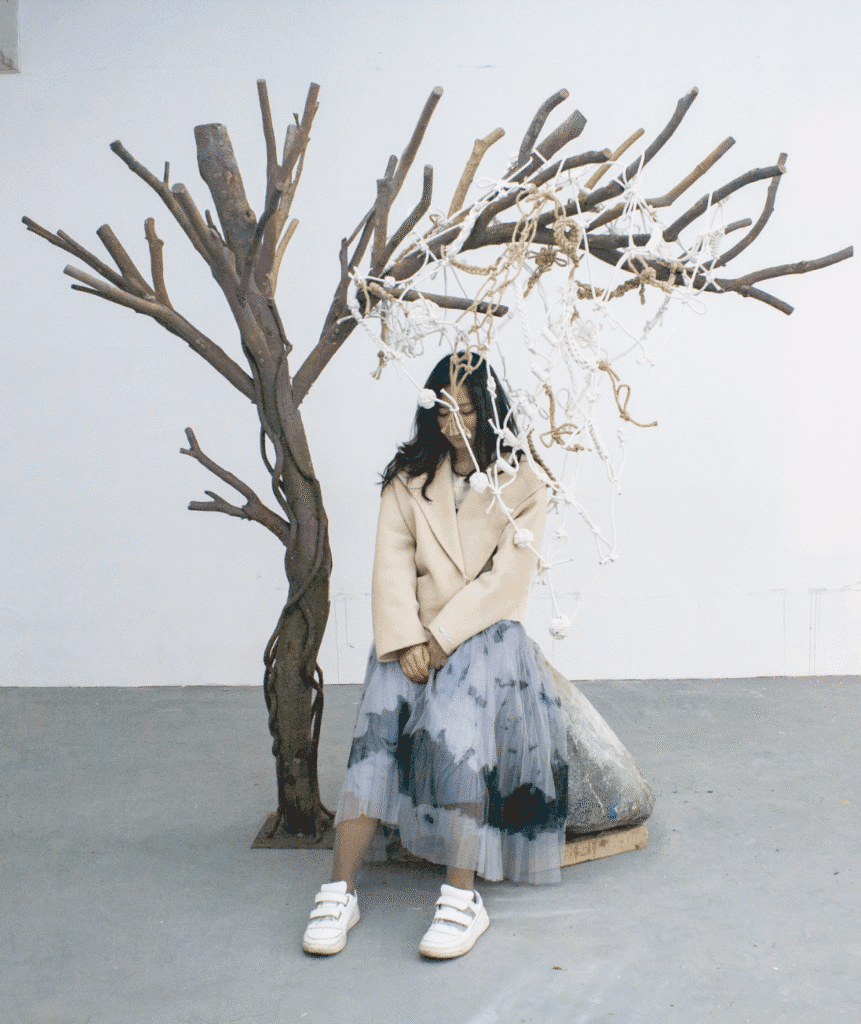 I sat under a knotted rope “tree” installation beside a stone with handprints, evoking Beckett’s Waiting for Godot and the existential act of waiting. CN: 艺术家安静坐在结绳“树”装置下,旁边是带手印的石头,呼应贝克特《等待戈多》,呈现等待的存在主义动作。