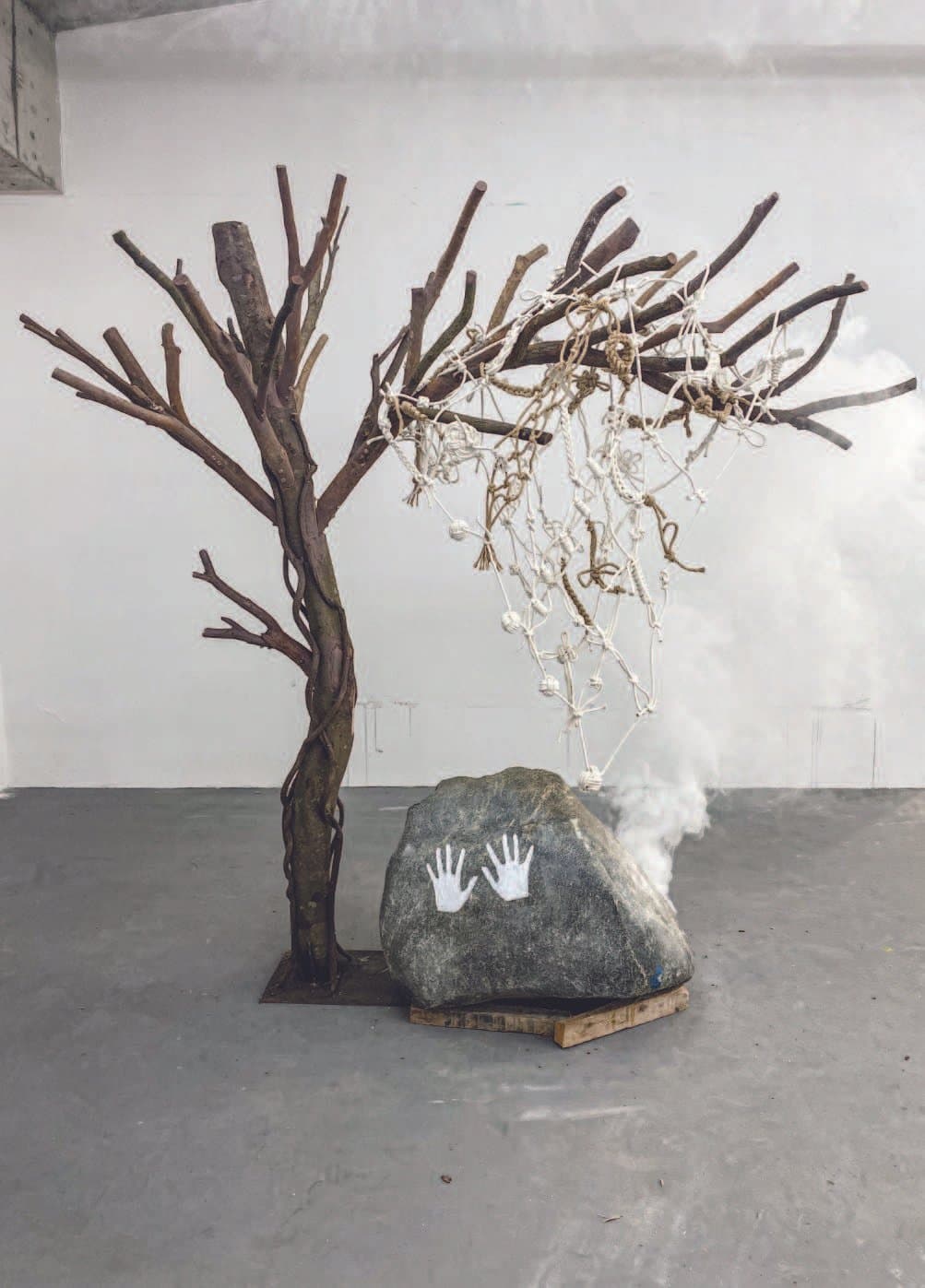 Installation of branch-built tree with sagging knotted rope net, a boulder marked by handprints, and drifting smoke — reflecting on the fragility of time and the necessity of living fully rather than merely recording.