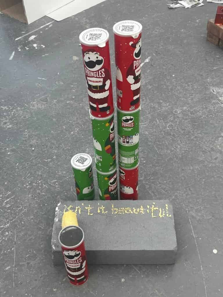Red and green Pringles cans stacked as towers beside crisps spelling Isn’t It Beautiful on a grey plinth.