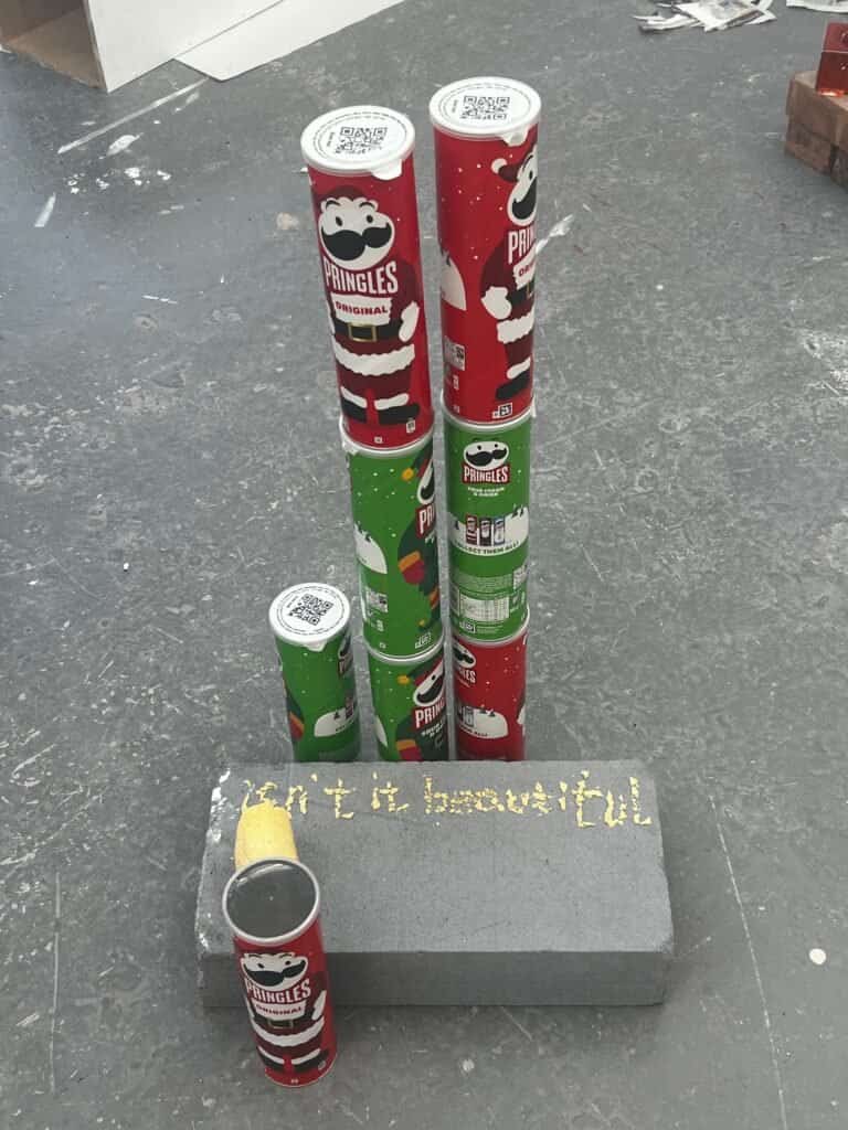 Red and green Pringles cans stacked as towers beside crisps spelling Isn’t It Beautiful on a grey plinth.