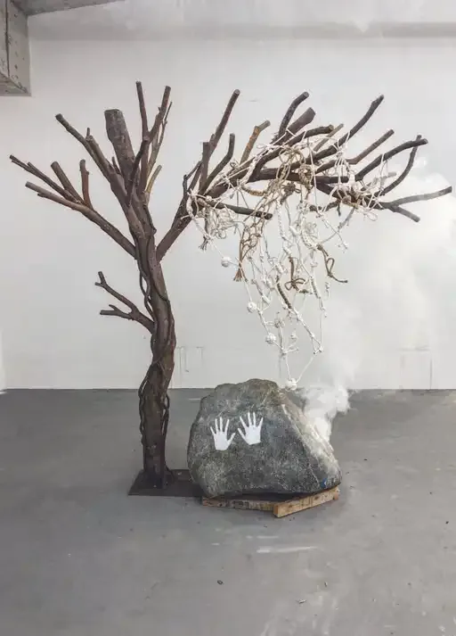 Installation of branch-built tree with sagging knotted rope net, a boulder marked by handprints, and drifting smoke — reflecting on the fragility of time and the necessity of living fully rather than merely recording.