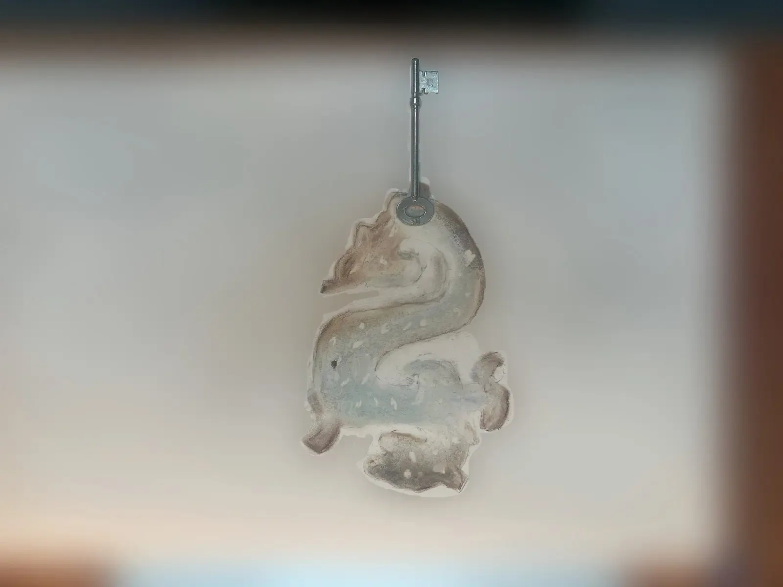 A pale translucent, heraldic-looking sculptural form hangs from an old metal key on a slender stand against a softly lit background.