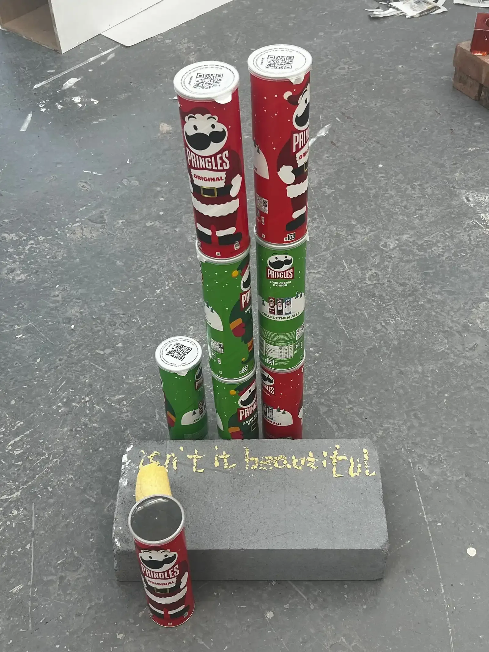 Red and green Pringles cans stacked as towers beside crisps spelling Isn’t It Beautiful on a grey plinth.