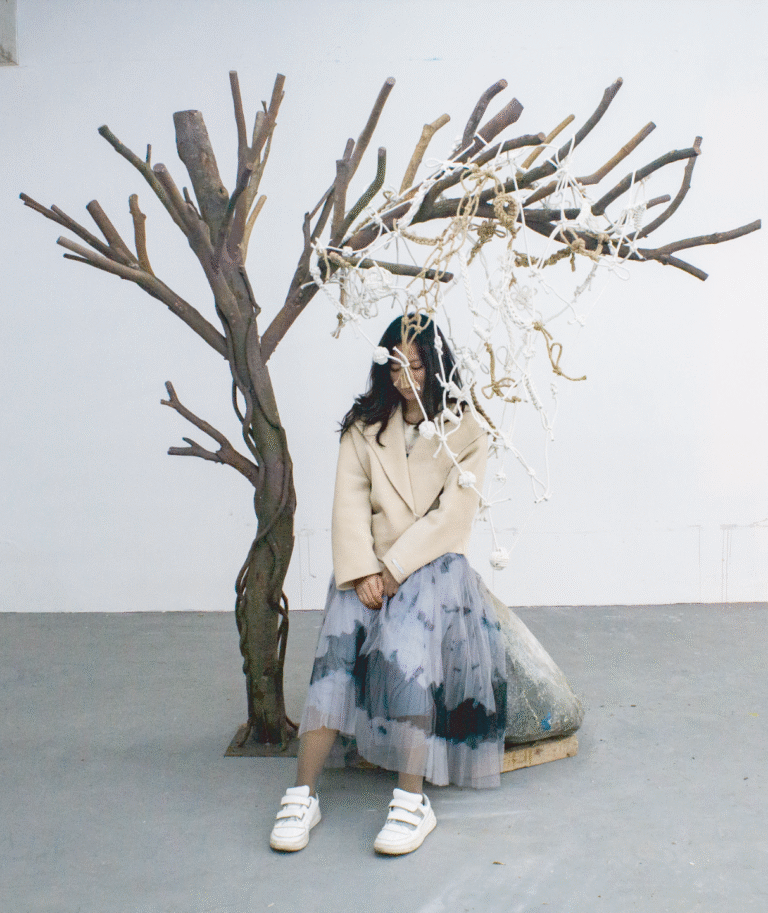 I sat under a knotted rope “tree” installation beside a stone with handprints, evoking Beckett’s Waiting for Godot and the existential act of waiting. CN: 艺术家安静坐在结绳“树”装置下,旁边是带手印的石头,呼应贝克特《等待戈多》,呈现等待的存在主义动作。