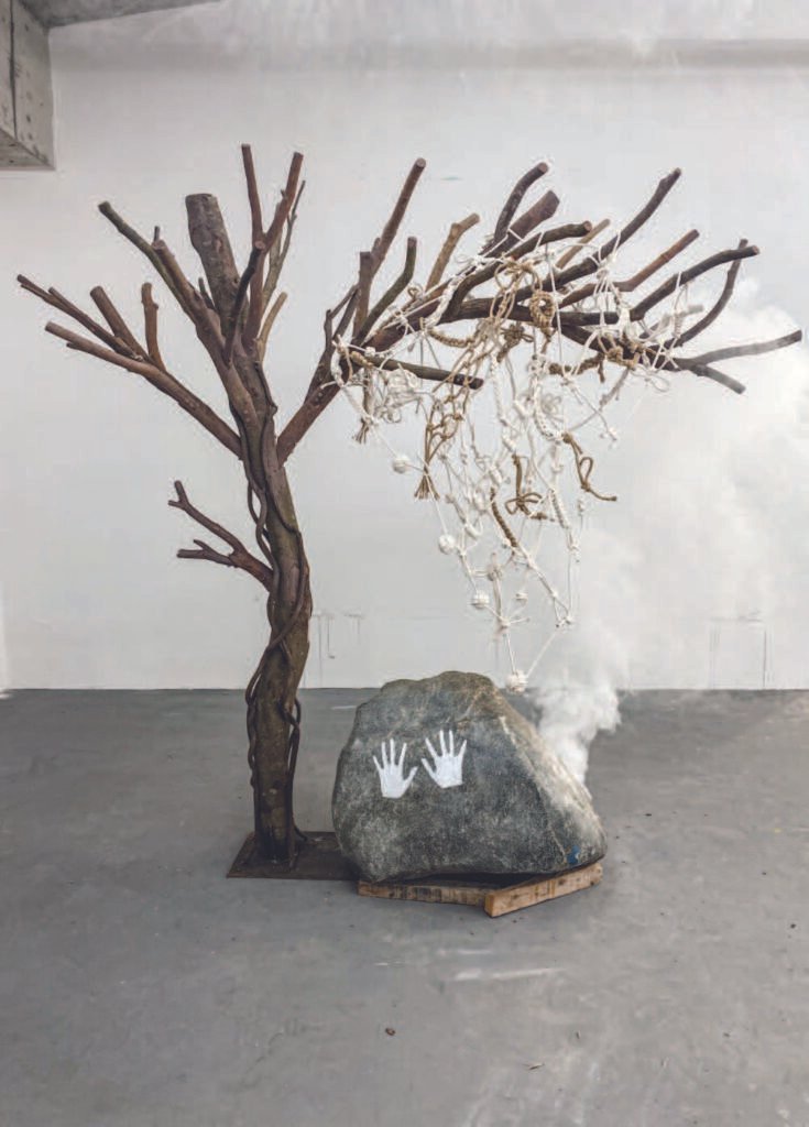 Installation of branch-built tree with sagging knotted rope net, a boulder marked by handprints, and drifting smoke — reflecting on the fragility of time and the necessity of living fully rather than merely recording.